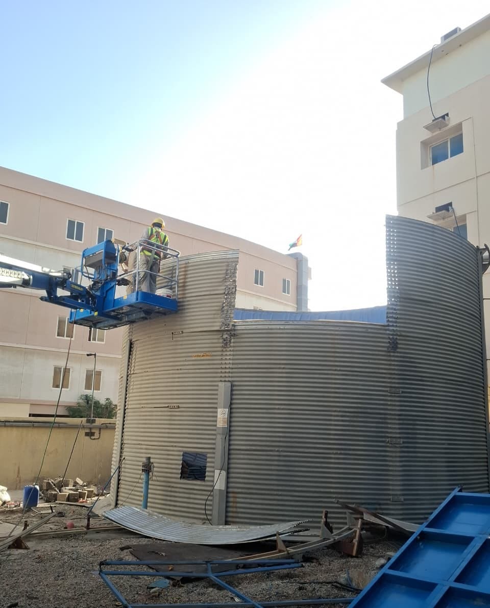 Worker on aerial platform cutting curved steel tank wall with debris being lowered safely at Jebel Ali